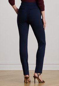 Žena oblečená vo slim-fit námorníckych nohaviciach a hnedých vysokých podpätkoch stojí na drevenej podlahe pred jednoducho stenou.