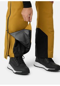 Waterdichte gele broek met zwarte accenten, voorzien van een ritszak bij de enkel, en zwarte laarzen met witte zolen.