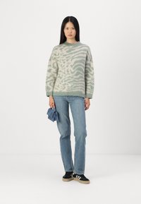 Maglione lavorato a maglia in verde chiaro e crema con un motivo ondulato, abbinato a jeans blu e scarpe nere con strisce bianche. Tiene in mano una piccola borsa blu.