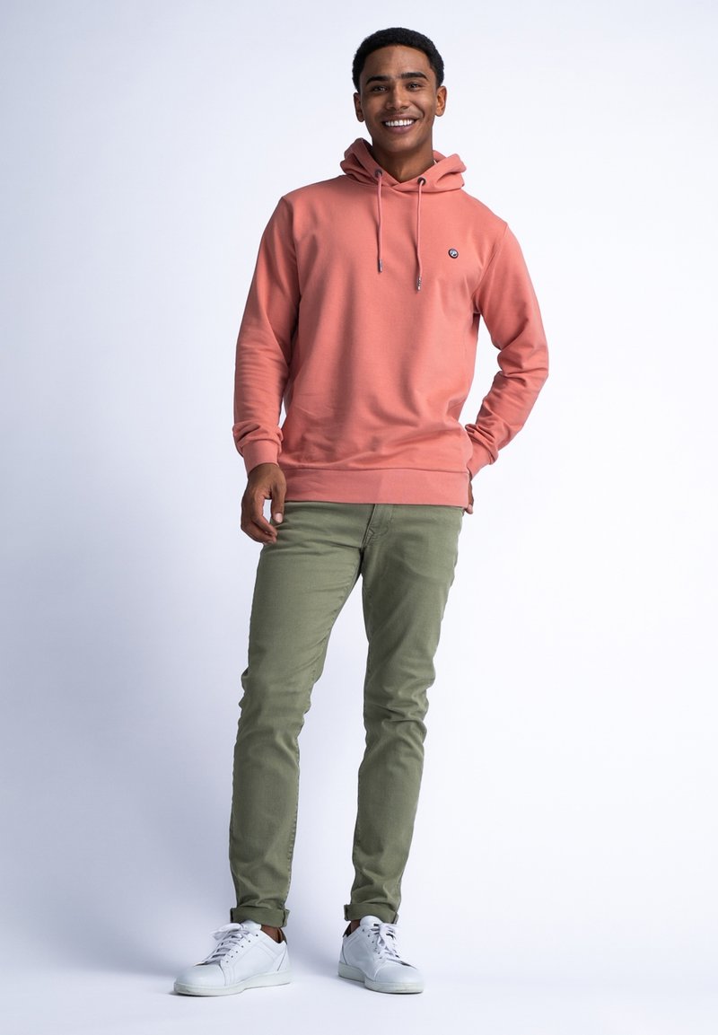 Sudadera de color rosa claro con cordón, pequeño logo, combinada con pantalones verdes oliva y zapatillas blancas sobre un fondo neutro.