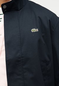 Κοντινή λήψη ναυτικού μπουφάν με το λογότυπο κροκόδειλου Lacoste στο στήθος πάνω από ροζ πουκάμισο και πράσινο ριγέ τοπ.