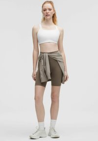 Baltas sportinis liemenėlė, rudos spalvos aukštu juosmeniu šortai, šviesiai smėlio spalvos džemperis, surištas aplink liemenį, derinamas su baltomis bateliais ir baltais kojiniais. Lygūs paviršiai.