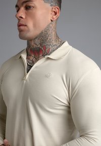 Muškarac s velikom tetovažom cvijeća na vratu i piercingom nosi bež majicu s patentnim zatvaračem dugih rukava i slojevite srebrne ogrlice na sivom backgroundu.