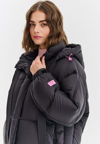 Kobieta o falowanych brązowych włosach, ubrana w czarną kurtkę puchową z różowymi akcentami, trzymająca kołnierz, na jednolitym jasnym tle.