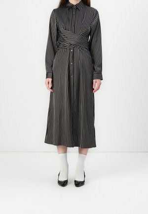 Donna che indossa un vestito nero a maniche lunghe con bottoni, righe verticali bianche e dettaglio a vita incrociata, abbinato a calzini bianchi e scarpe nere.