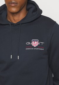 Gros plan sur une personne portant un sweat à capuche bleu marine avec le logo "GANT 1949" et "American Sportswear" brodé sur la poitrine.