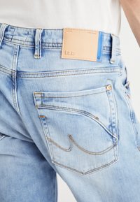 Jeans di denim azzurro chiaro presentano una toppa in pelle beige sulla vita, cuciture gialle e due tasche posteriori con dettagli a cucitura curva.