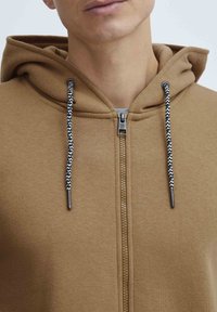 Haut à capuche zippé marron en tissu texturé, avec des cordons de serrage à motifs noir et blanc et une fermeture éclair à l'avant.