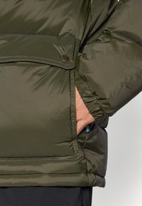 Veste rembourrée vert olive avec une texture lisse et brillante, dotée d'une fermeture à boutons-pression et d'un poignet ajustable. Poche visible avec une étiquette.