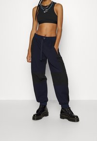 Žena v černém zkráceném topu Nike, tmavě modrých a černých džínových joggerech s nášivkami, černých platformových botách na šňůrky a s vrstvenými korálkovými náhrdelníky.