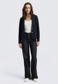 Navy blazer with two buttons, paired with a white top and high-waisted dark jeans. Model wears white sneakers, standing on a neutral background.