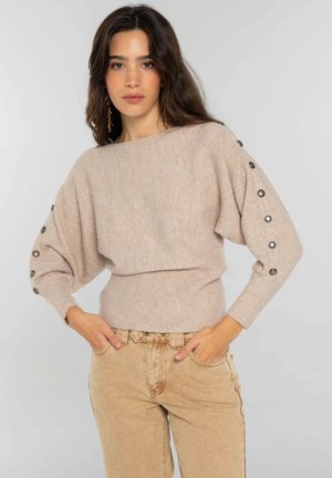 Suéter beige de punto con cuello amplio, mangas abullonadas y botones decorativos a lo largo del puño. Combinado con pantalones de tiro alto en marrón claro.