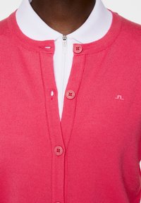 Cardigan lavorato a maglia rosa con colletto rotondo, chiusura frontale con zip e due bottoni in tinta. Sotto una camicia bianca con colletto, caratterizzata da un ricamo del logo discreto.