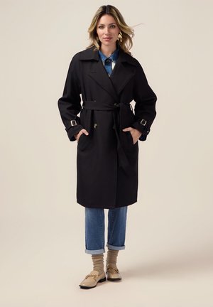 Zwarte trenchcoat met een ceintuur in de taille, dubbele knopen aan de voorkant en zijzakken. Gecombineerd met omgeslagen spijkerbroeken en beige instappers.