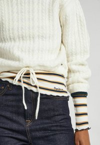 Maglione bianco lavorato a maglia con texture a costine e coulisse in vita, caratterizzato da un orlo a strisce nei colori blu navy, arancione e crema. Indossato con jeans scuri.