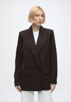 Lyshåret kvinde iført oversized dobbeltbrystet sort blazer over hvid skjorte og hvide bukser, stående mod en ensfarvet lys baggrund.