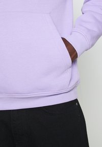 Levandulová mikina s přední kapsou a žebrovanými manžetami, kombinovaná s černými kalhotami, vyznačující se hladkou texturou a ležérním designem.