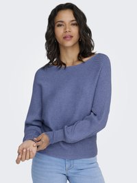 Frau mit welligem dunklem Haar, die einen blauen, gerippten Pullover und hellblaue Jeans trägt, und den Ärmel vor einem hellgrauen Hintergrund hochkrempelt.