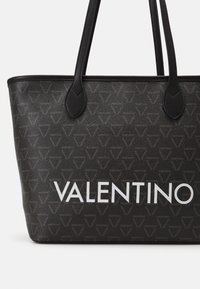 Svart tote-väska i texturerat material, med ett geometriskt mönster och vit "VALENTINO"-text på framsidan. Dubbla svarta handtag.