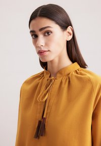 Senfgelbe Bluse mit hohem Kragen, minimalen Falten und einer Krawatte mit Quasten. Aus glattem Stoff gefertigt und mit einer lockeren Passform. 