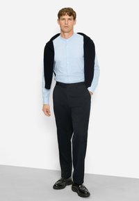 Helles blaues Hemd mit Stehkragen, kombiniert mit schwarzen Hosen und einem dunklen Pullover, der über die Schultern drapiert ist. Schwarze Schuhe runden den Look ab.