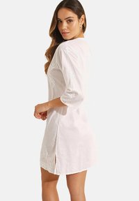 Witte linnen jurk met 3/4 mouwen, voorzien van bloemenborduurwerk en zijsplitten. Zachte textuur, comfortabele pasvorm en ronde onderrand.