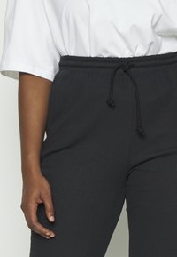 Henkilö, joka on pukeutunut mustiin ribattuihin jogger-housuihin, joissa on kiristysnauha, ja valkoiseen lyhythihaiseen paitaan, käsi lepää lähettyvillä reidellä.