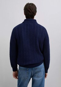 Pull en maille torsadée bleu marine avec un col haut et des poignets côtelés, présentant une texture épaisse et une coupe décontractée. Porté avec un jean bleu clair.