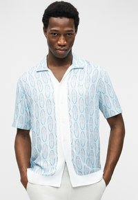 Νέος άνδρας με τα χέρια στις τσέπες, φορώντας ανοιχτό μπλε πουκάμισο με μοτίβο και κοντά μανίκια πάνω από λευκό πουκάμισο με κουμπιά, μπροστά σε απλό φόντο.