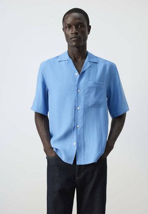 Hombre vestido con camisa azul clara de manga corta con botones y bolsillo en el pecho, y pantalones oscuros, de pie con las manos en los bolsillos contra un fondo liso.