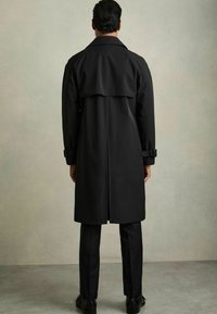 Pardesiu negru cu guler înalt, cupe cu curea și detalii de capă pe spate. Purtați peste o pereche de pantaloni închiși și pantofi negri lucioși. Textură netedă.