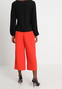 Musta pitkähihainen toppi, jossa on pleatatut hihat, yhdistetty kirkkaanpunaisiin culotteihin. Malli mustissa korkokengissä, selkä kameraan päin.