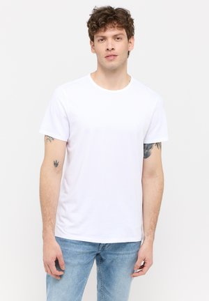 Tricou din bumbac alb, cu mâneci scurte, decolteu rotund, stil mulat. Prezintă un detaliu subtil cu logo pe piept. Asortat cu blugi de un albastru deschis.
