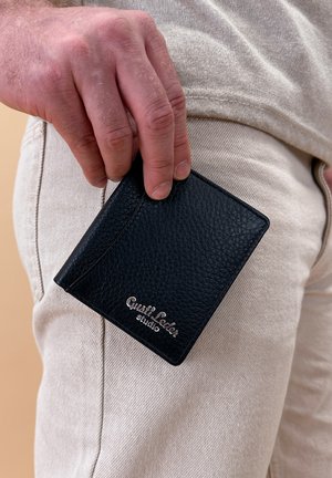 Schwarze Lederbörse mit einer genarbten Textur, die ein elegantes Design und ein geprägtes Logo aufweist. Auf hellen Hosen platziert.