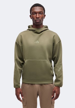 Άνδρας ντυμένος με ελιάς πράσινη κουκούλα Adidas και μπεζ παντελόνια στέκεται μπροστά σε απλό ανοιχτό υπόβαθρο, κοιτώντας μπροστά με ουδέτερη έκφραση.