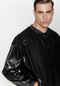 Czarna kurtka typu varsity z wełnianym korpusem, błyszczącymi czarnymi skórzanymi rękawami, okrągłym kołnierzem oraz haftem nuty muzycznej na klatce piersiowej.