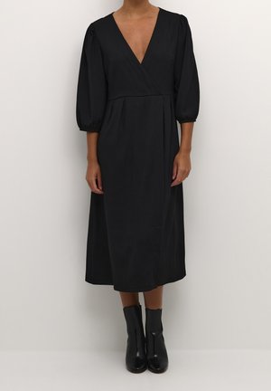 Woman wearing a black midi dress with three-quarter puff sleeves, deep V-neck, and black ankle boots against a plain background.