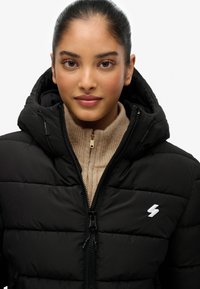 Sort puffjakke med lynlås fortil og hætte, der har en ribbet beige sweater underneath. Bemærkelsesværdig hvid logo-detalje på brystet.
