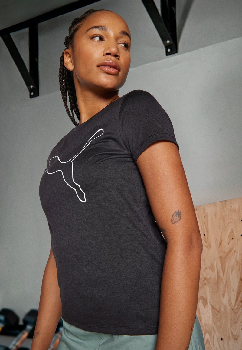 Tricou negru ajustat cu mâneci scurte, având un logo alb Puma pe partea din față. Textură netedă, purtat cu pantaloni de culoare verde deschis.