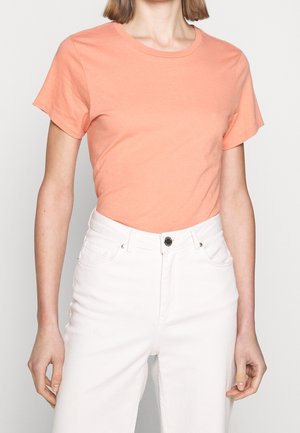 Personne portant un t-shirt pêche à manches courtes avec col rond, rentré dans un jean blanc taille haute avec boutons et poches visibles.