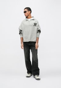 Siv pleten pulover s črnimi poudarki, z ovratnikom, rebrastim spodnjim robom. Črne široke kavbojke z diskretnimi detajli. Sivi in beli športni copati, temna sončna očala.