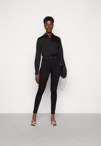 Schwarzes Hemd mit Knopfleiste, hochgeschnittene schwarze Skinny-Jeans, weiße Riemchen-Absätze und eine schwarze Clutch mit einer silbernen Kette. Glatte Textur insgesamt.