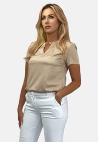 Beżowa bluzka z krótkim rękawem i dekoltem w serek z metalicznym akcentem. Połączona z białymi spodniami o dopasowanym kroju, widoczne kieszenie, gładka tekstura materiału.