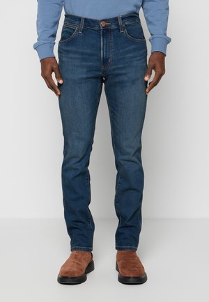 Mężczyzna w dopasowanych niebieskich dżinsach, jasnoniebieskiej bluzie i brązowych zamszowych butach, stojący na jednolitym szarym tle.