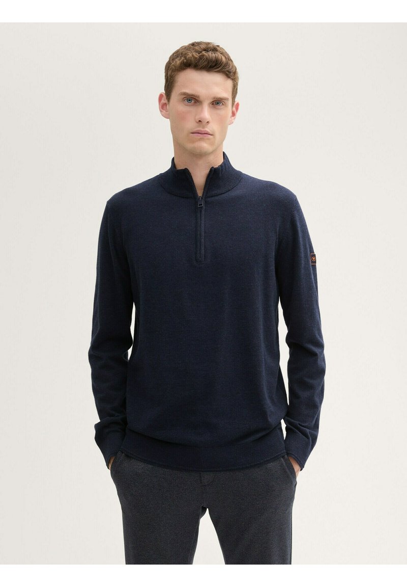 Marinefarbener Reißverschluss-Pullover aus weichem Material, mit langen Ärmeln, Stehkragen und geripptem Saum. Kleines Logo-Detail am Ärmel.