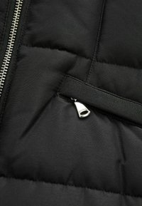 Sección de chaqueta acolchada negra mostrando una textura acolchada, detalle de cremallera plateada y un tirador de cremallera de tela negra en contraste.