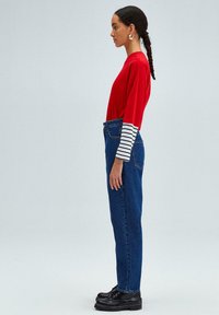 Camiseta roja de manga larga con puños a rayas en blanco y negro, combinada con jeans de mezclilla azul de cintura alta y botas negras con cordones.