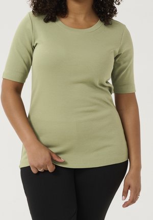 Woman with curly hair wearing a short-sleeve, light green fitted ribbed shirt and black pants against a plain background.