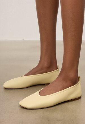 Jambes bronzées portant des ballerines en cuir jaune pâle à bout carré sur un sol beige lisse.