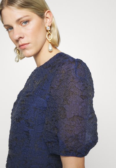 Vestido azul marinho com padrão floral, confeccionado com tecido texturizado, apresentando mangas bufantes e decote redondo. Brincos dourados com detalhes em forma de gota.